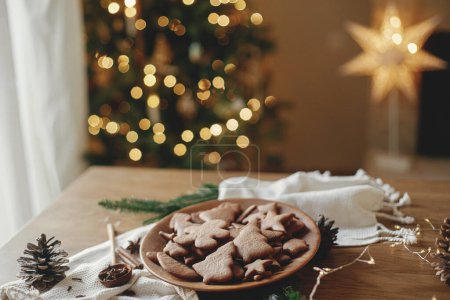 Foto de Deliciosas galletas de jengibre en plato de madera sobre mesa rústica contra elegantes luces de árbol de Navidad bokeh. Galletas de pan de jengibre de Navidad recién horneadas, tiempo de vacaciones atmosféricas - Imagen libre de derechos
