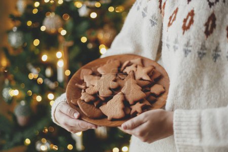 Photo for Christmas gingerbread cookies in wooden plate in hands on background of stylish christmas golden illumination. Merry Christmas! Delicious gingerbread cookies, atmospheric holiday - Royalty Free Image