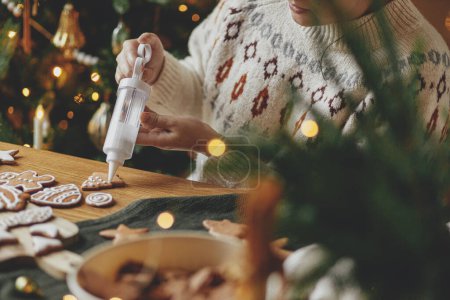 Foto de Decorar galletas de jengibre con hielo en la mesa de madera rústica en las luces doradas del árbol de Navidad. Tradiciones navideñas atmosféricas. Mujer decorando galletas con glaseado de azúcar - Imagen libre de derechos