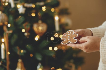 Photo for Hand holding christmas gingerbrea man cookie with icing against festive christmas tree with golden lights bokeh. Merry Christmas! Delicious gingerbread cookies, atmospheric holiday time - Royalty Free Image