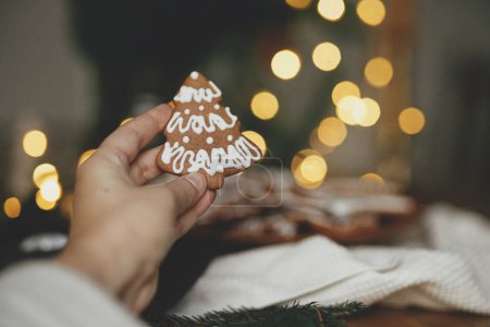 Téléchargez les photos : Main tenant Noël pain d'épice biscuit d'arbre avec glaçage contre les lumières dorées festives bokeh. Joyeux Noël ! Délicieux biscuits au pain d'épice, temps des fêtes atmosphériques - en image libre de droit