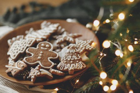 Photo for Merry Christmas! Gingerbread cookies with icing in plate on wooden rustic table with fir branches, festive decorations and christmas golden lights. Atmospheric Christmas holidays, family time - Royalty Free Image