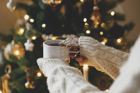 Foto de Manos en suéter sosteniendo cálida taza de té con galletas de jengibre de Navidad contra el árbol de Navidad festivo con luces doradas bokeh. ¡Feliz Navidad! Tiempo de vacaciones atmosféricas, hygge de invierno - Imagen libre de derechos