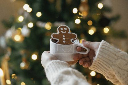 Foto de ¡Feliz Navidad! Manos en suéter sosteniendo taza de té caliente con galleta de jengibre hombre contra el árbol de Navidad con luces doradas bokeh. Vacaciones de Navidad atmosféricas, hygge de invierno - Imagen libre de derechos