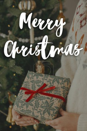 Photo for Merry Christmas text on stylish christmas gift with red ribbon in hands close up at modern decorated vintage tree. Happy Holidays! Season's greeting card. Handwritten sign - Royalty Free Image