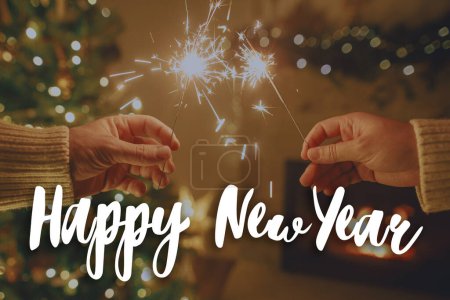 Photo for Happy New Year text on burning sparklers in hands on background of  fireplace and christmas tree lights. Fireworks glowing, festive celebration. Season's greeting card. Handwritten sign - Royalty Free Image