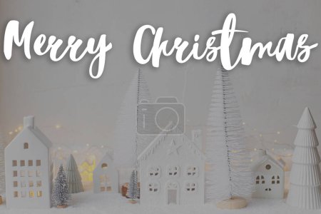Photo for Merry Christmas text on stylish little white houses and trees, snowy christmas miniature village  with golden lights. Happy Holidays! Season's greeting card. Handwritten sign - Royalty Free Image