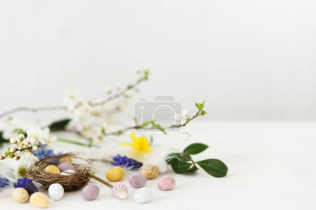 Photo for Happy Easter! Stylish easter chocolate eggs in nest, spring flowers, feathers border on white rustic wooden table. Easter modern simple decoration banner, space for text. Seasons greetings - Royalty Free Image