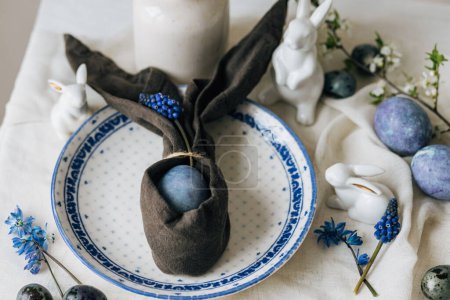 Foto de ¡Feliz Pascua! Elegante huevo de Pascua en servilleta de conejo en plato vintage y flores de primavera en servilleta de lino, ajuste de mesa rústico. Huevos azules pintados naturales y flores de jacinto. Moderno bodegón mínimo - Imagen libre de derechos
