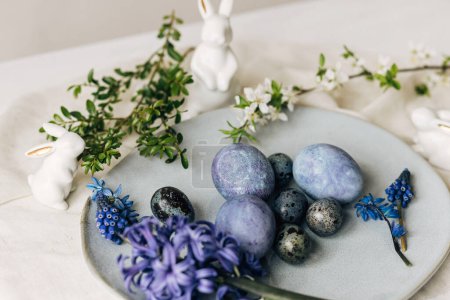 Foto de Elegantes huevos de Pascua en plato vintage, flores de conejo y primavera en mesa rústica. ¡Feliz Pascua! Huevos azules tinte natural, flores hiacynt púrpura en servilleta de lino. Ambiente de vacaciones, naturaleza muerta mínima - Imagen libre de derechos
