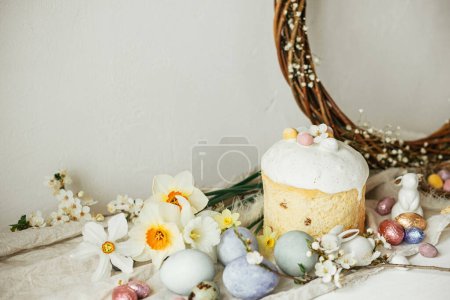 Stylish easter eggs, panettone, bunnies, cherry blossom and daffodils composition on rustic table. Happy Easter! Modern natural dyed eggs, festive food and spring flowers still life
