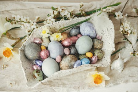 Téléchargez les photos : Joyeux Pâques ! Des œufs de Pâques élégants dans un bol moderne avec des fleurs de cerisier et des jonquilles sur une table rustique. Oeufs teints naturels modernes et fleurs de printemps. Pâques nature morte décor dans la maison de campagne - en image libre de droit