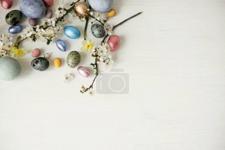 Téléchargez les photos : Des œufs de Pâques élégants et des fleurs de cerisier à plat reposent sur une table blanche rustique. Joyeux Pâques ! Oeufs teints naturels modernes et en chocolat avec des fleurs de printemps. Modèle festif avec espace de copie - en image libre de droit