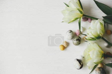 Foto de Elegantes huevos de Pascua y tulipanes sobre una mesa blanca rústica. ¡Feliz Pascua! Pascuas planas. Moderno colorante natural de mármol y dulces huevos de chocolate con borde de flores de primavera. Espacio para texto - Imagen libre de derechos