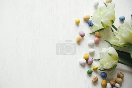 Foto de ¡Feliz Pascua! Ponen con estilo los huevos de Pascua y los tulipanes sobre la mesa rústica blanca plana. Huevos naturales de chocolate con flores de primavera. Frontera festiva de Pascua, espacio para texto - Imagen libre de derechos