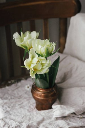 Foto de Hermosos tulipanes blancos en jarrón vintage en silla de madera vieja con composición de tela de lino. Naturaleza muerta en primavera. Flores de primavera en habitación rústica moody - Imagen libre de derechos