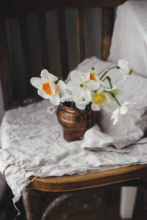 Photo for Stylish daffodils in vintage vase on old wooden chair with linen cloth.  Rustic still life. Countryside moody spring flowers composition. Easter floral banner - Royalty Free Image