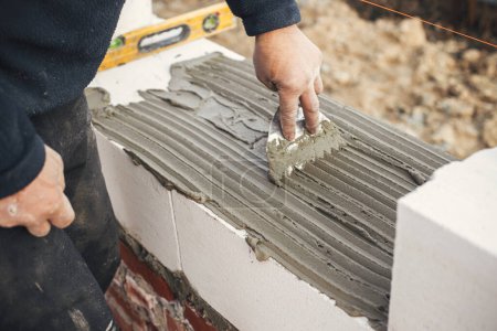 Foto de Worker laying autoclaved aerated concrete blocks, working with adhesive and trowel. Builder installing masonry white  blocks close up. Process of house building at construction site - Imagen libre de derechos