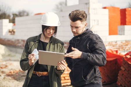 Photo for Stylish woman architect with tablet  and contractor man checking blueprints at construction site. Young engineer or construction workers in hardhat looking at plans of new modern house - Royalty Free Image