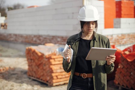 Téléchargez les photos : Stylish woman architect with tablet checking blueprints at construction site. Young female engineer or construction worker in hardhat looking at plans of new modern house - en image libre de droit