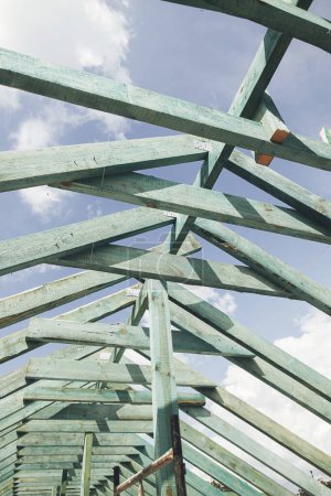 Foto de Unfinished roof trusses, view in attic with rafters and beams against sky. Wooden roof framing. Mansard of modern farmhouse building construction. - Imagen libre de derechos