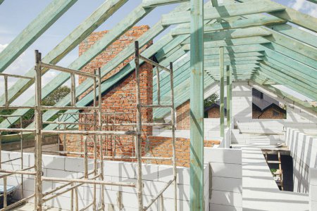 Foto de Unfinished roof trusses, view in attic with rafters, beams, windows and chimney. Wooden roof framing. Mansard of modern farmhouse building construction. - Imagen libre de derechos