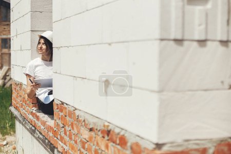 Foto de Joven arquitecta con tableta revisando planos mientras está sentada en la ventana de la nueva casa moderna. Ingeniera mujer con estilo en el sombrero duro mirando los planes digitales en la tableta en el sitio de construcción - Imagen libre de derechos
