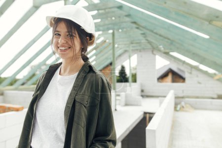 Photo for Stylish woman architect or engineer smiling against wooden roof framing of modern farmhouse. Portrait of young happy female in hard hat at construction site. Copy space - Royalty Free Image