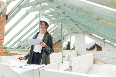 Photo for Young happy female architect with tablet checking blueprints against wooden roof framing of modern farmhouse. Stylish woman engineer in hard hat looking at digital plans at construction site - Royalty Free Image