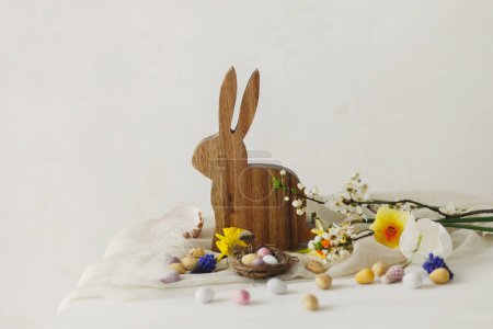 Photo for Happy Easter! Stylish easter chocolate eggs in nest, spring flowers, bunny figurine on white rustic wooden table. Easter modern simple banner, space for text. Seasons greetings - Royalty Free Image