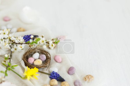 Téléchargez les photos : Œufs de Pâques colorés en chocolat dans le nid, fleurs de printemps, plumes et composition de bordure en lin sur table en bois blanc. Espace pour le texte. Joyeux Pâques ! Salutations saisonnières - en image libre de droit
