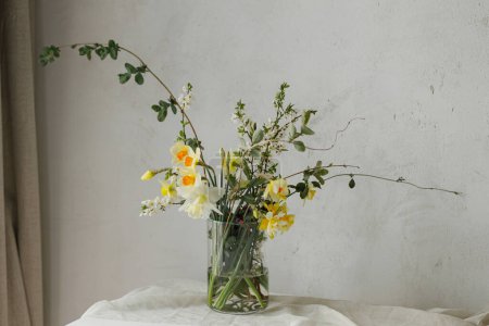 Foto de Elegante ramo de flores de primavera en la mesa rústica en la habitación rural. Hermosos narcisos, flor de cerezo y composición de vegetación en jarrón de vidrio. Pascua decoración sencilla moderna - Imagen libre de derechos