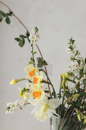 Photo for Stylish spring flowers bouquet on rustic table in rural room. Beautiful daffodils, cherry bloom and greenery composition in glass vase. Easter modern simple decor - Royalty Free Image