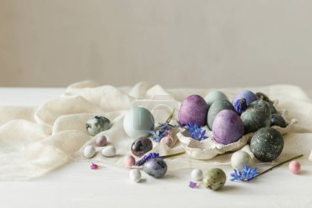 Foto de Elegantes huevos de Pascua en bandeja moderna, suaves flores de primavera y tela de lino sobre una mesa blanca rústica. ¡Feliz Pascua! Mármol pintado natural y huevos morados bodegón. Saludos de temporada - Imagen libre de derechos