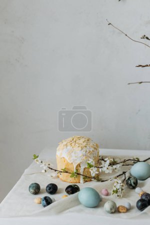 Stylish easter eggs, panettone and spring flowers on linen cloth on rustic table. Natural dye eggs and cherry blossom, festive minimal still life. Happy Easter!