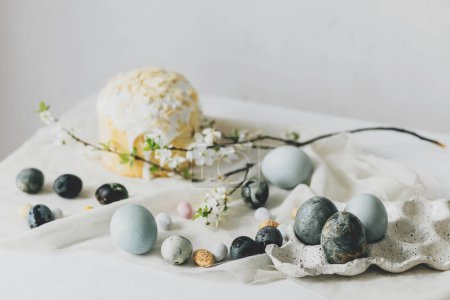 ¡Feliz Pascua! Elegantes huevos de Pascua, pan casero de Pascua y flores de primavera en la servilleta de lino en la mesa rústica contra la pared. Natural pintado azul y huevos de mármol y flores de cerezo bodegón