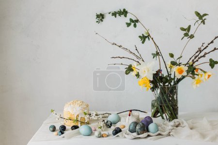 Stilvolle Ostereier, Panettone und Frühlingsblumen auf Leinentuch auf rustikalem Tisch. Natürliche Farbstoffe Eier und Narzissen Bouquet, festliche minimale Stillleben. Frohe Ostern!
