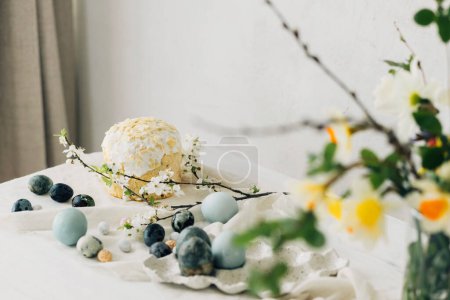Happy Easter! Stylish easter eggs, homemade easter bread and spring flowers on linen napkin on rustic table against wall. Natural painted blue and marble eggs and cherry blooms still life