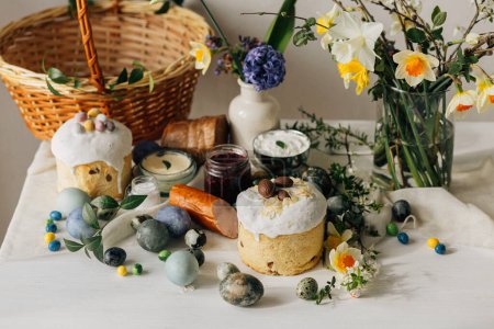 Elegantes huevos de Pascua, pan y cesta con flores de primavera en la mesa rústica. ¡Feliz Pascua! Comida tradicional de Pascua para bendecir. Huevos de tinte naturales modernos, jamón sabroso, pan, mantequilla, remolacha