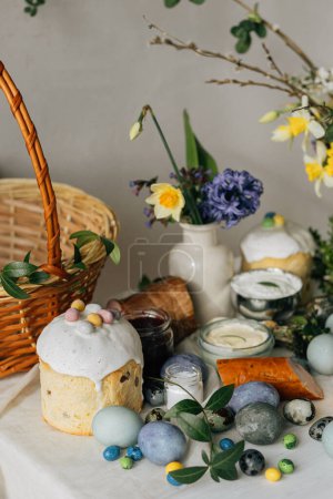 Foto de Elegantes huevos de Pascua, pan y cesta con flores de primavera en la mesa rústica. ¡Feliz Pascua! Comida tradicional de Pascua para bendecir. Huevos de tinte naturales modernos, jamón sabroso, pan, mantequilla, remolacha - Imagen libre de derechos