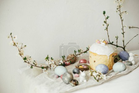 Foto de Elegantes huevos de Pascua, panettone, conejos, composición de flores de cerezo en la mesa rústica. ¡Feliz Pascua! Huevos teñidos naturales modernos, comida festiva y flores de primavera. Naturaleza muerta rural - Imagen libre de derechos