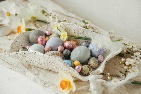 Photo for Stylish easter eggs in modern bowl with cherry blossom and daffodils composition on rustic table. Happy Easter! Modern colorful natural dyed eggs, chocolate and spring flowers. Easter hunt - Royalty Free Image