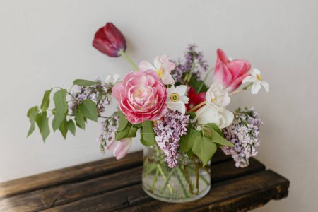 Foto de Elegante ramo de primavera en un banco de madera rústico. Hermosas lilas, tulipanes y narcisos en jarrón de cristal sobre fondo de pared gris. Bodegón floral. Feliz Día de las Madres - Imagen libre de derechos