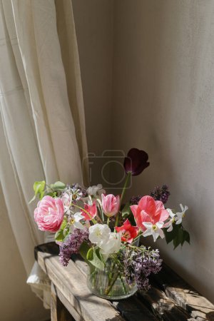 Téléchargez les photos : Bouquet de printemps élégant sur banc en bois rustique. Beaux lilas, tulipes et jonquilles en vase de verre sur fond de mur gris et rideaux de lin. Nature morte florale. Bonne fête des mères - en image libre de droit