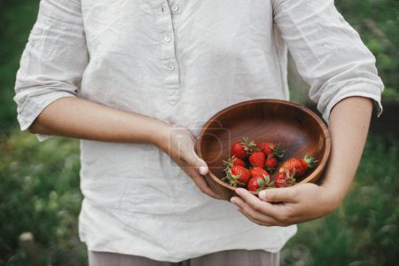 Téléchargez les photos : Femme tenant un bol en bois avec des fraises fermer. Récolte de baies naturelles fraîches dans un jardin bio urbain. Style de vie Homestead - en image libre de droit