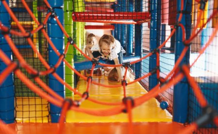 Happy group of siblings crawling and playing together in indoor playground. Excited kids playing together on net ropes. Cute school kids playing on the colorful playground