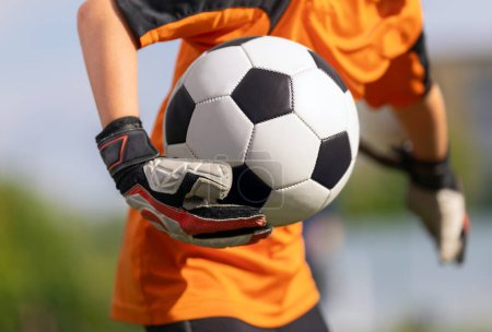 Téléchargez les photos : Young boy as a soccer goalie holding the ball in one hand ready to start a game. Football goalkeeper in jersey shirt and sports gloves play a football match - en image libre de droit