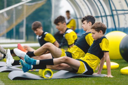 Teenage boys at football training in soccer club. Sports summer camp for youth footballers. Boys using foam therapy rollers after training session outdoor