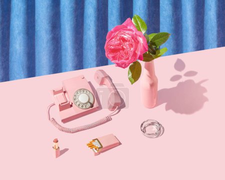 Retro vintage scene with old dial phone, shiny ash tray, rose flower and cigarette pack on bright pink table. Blue plush curtain background.