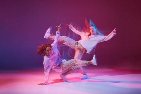 Foto de Retrato de chicas jóvenes bailando hip-hop aisladas sobre fondo violeta degradado en neón con luz mixta. Movimientos caóticos. Concepto de movimiento, cultura juvenil, estilo de vida activo, acción, danza callejera - Imagen libre de derechos